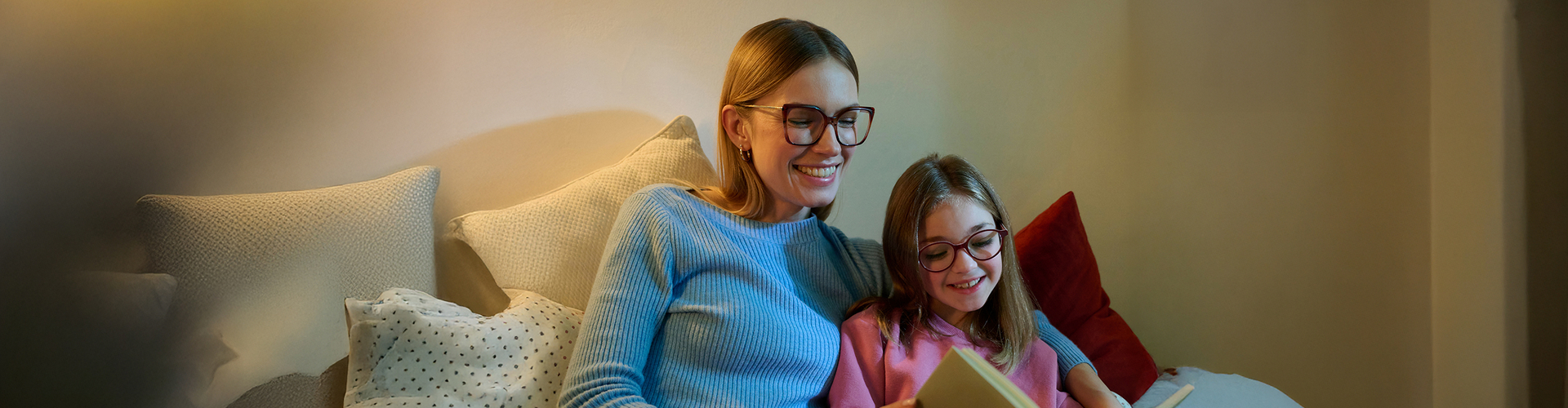 Mutter und Tochter sitzen gemütlich auf dem Sofa und lesen gemeinsam ein Buch, beide tragen Brillen und lächeln.