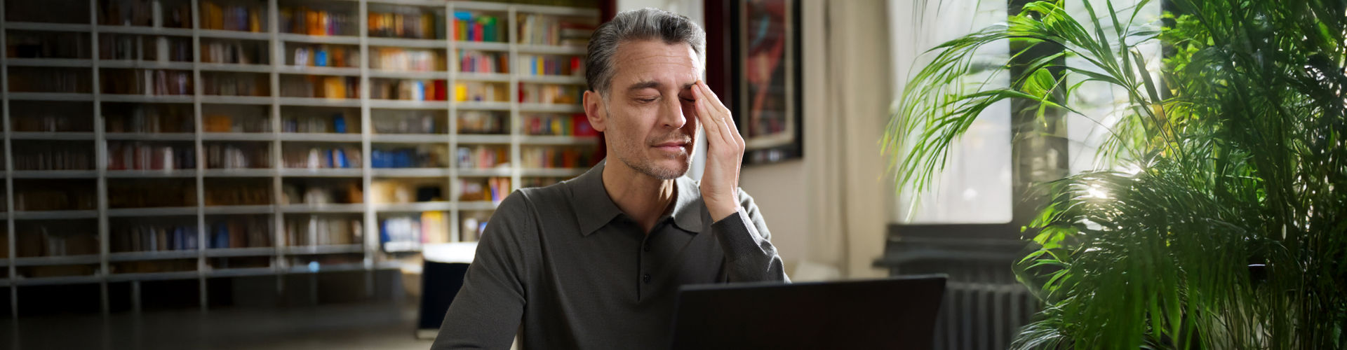 Ein Mann sitzt vor einem Bücherregal & arbeitet mit seinem Laptop. Währenddessen hält er seine Hand an sein Auge.