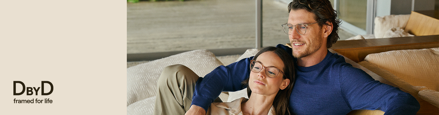Deux personnes assises ensemble sur un canapé, portant toutes deux des lunettes DbyD, dans une ambiance chaleureuse et détendue près d’une grande fenêtre.