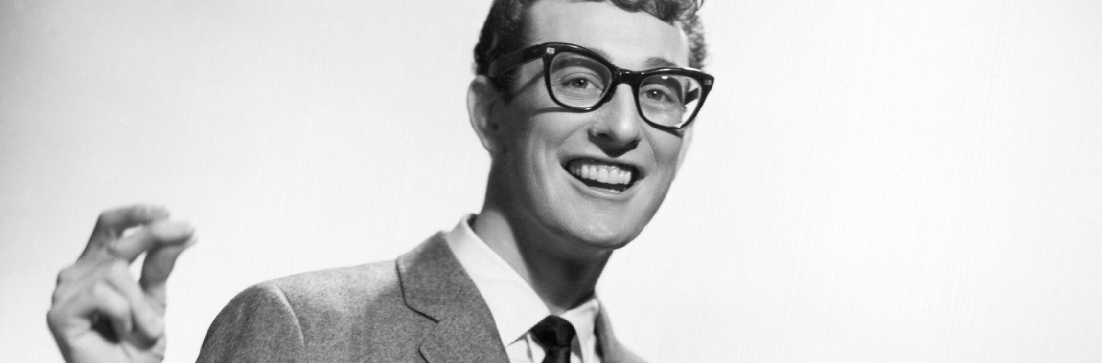 Black and white, head and shoulders photograph of Buddy Holly, wearing black glasses and a shirt and tie.