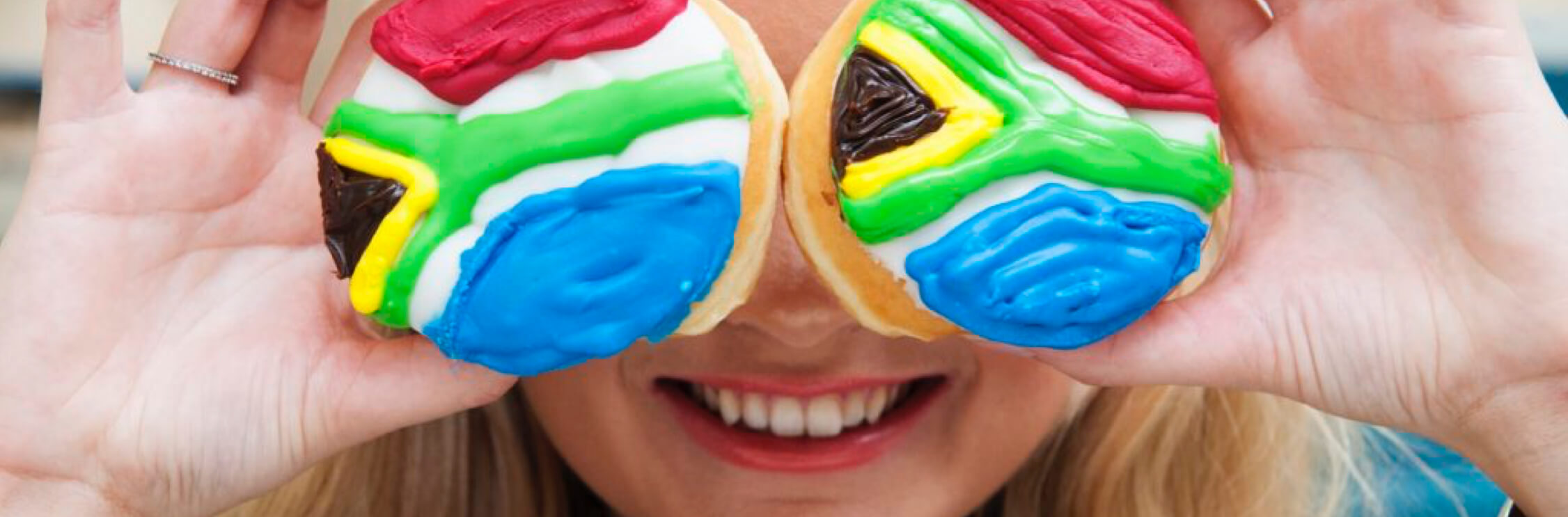Close up of young woman with blonde hair holding up to her eyes two sponge cakes decorated with the South African flag in icing.