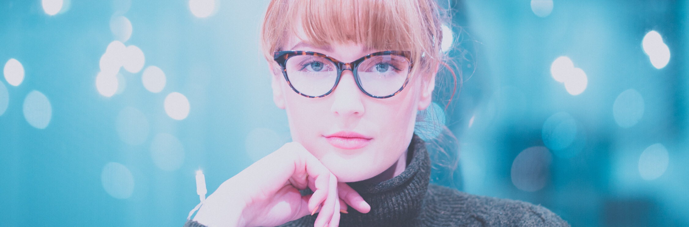 Head and shoulders image of a young woman with red hair against a blue background wearing a green turtle-neck jumper and large tortoiseshell cats-eye glasses.